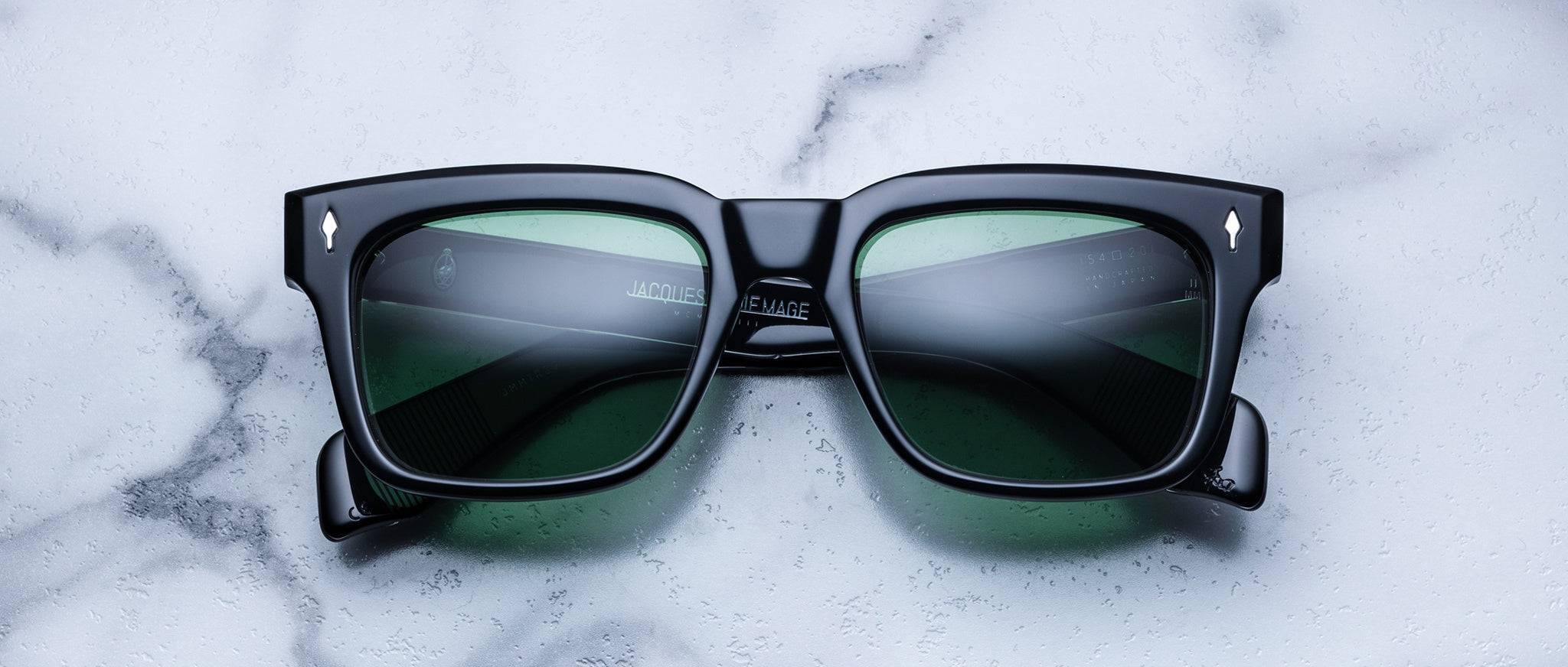 The Jacques Marie Mage Torino by Jacques Marie Mage features black bold rectangular acetate frames with green-tinted lenses, displayed on white marble and highlighting sophisticated handcrafted eyewear.
