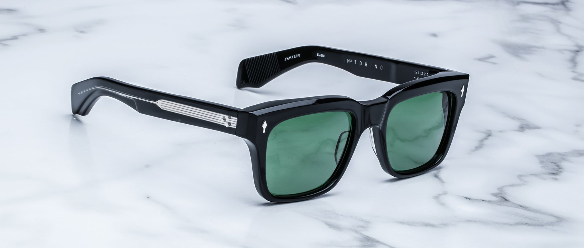 The Jacques Marie Mage Torino sunglasses by Jacques Marie Mage feature bold black rectangular acetate frames with green lenses, elegantly displayed on a white marble surface.