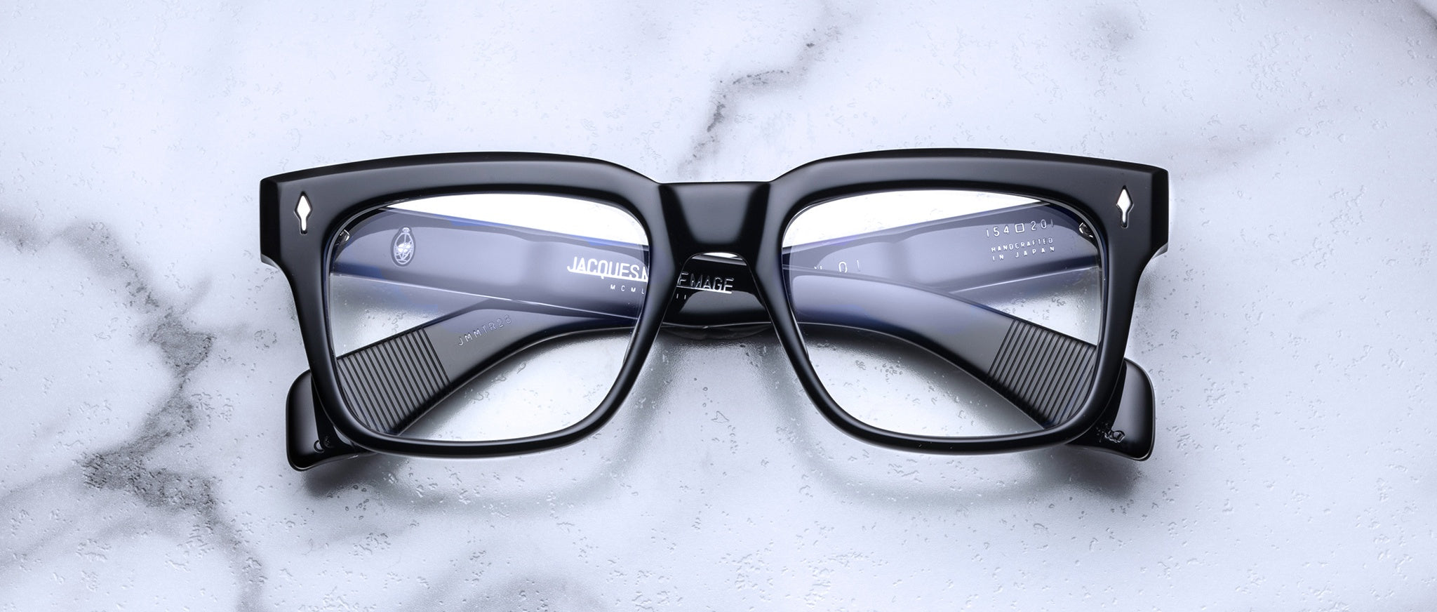 The Jacques Marie Mage Torino by Jacques Marie Mage features black rectangular frames made from handcrafted acetate, showcased on a white marble surface and viewed from above.