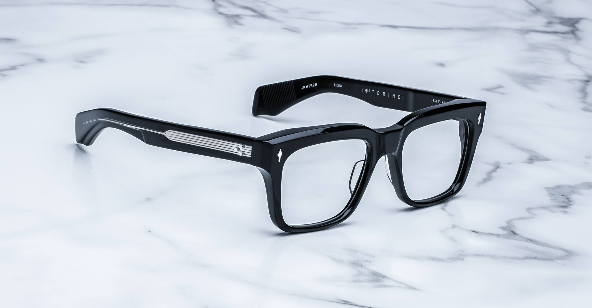 A pair of bold, handcrafted Jacques Marie Mage Torino rectangular acetate eyeglass frames is displayed on a white marble surface, showcasing the distinctive style of Jacques Marie Mage.