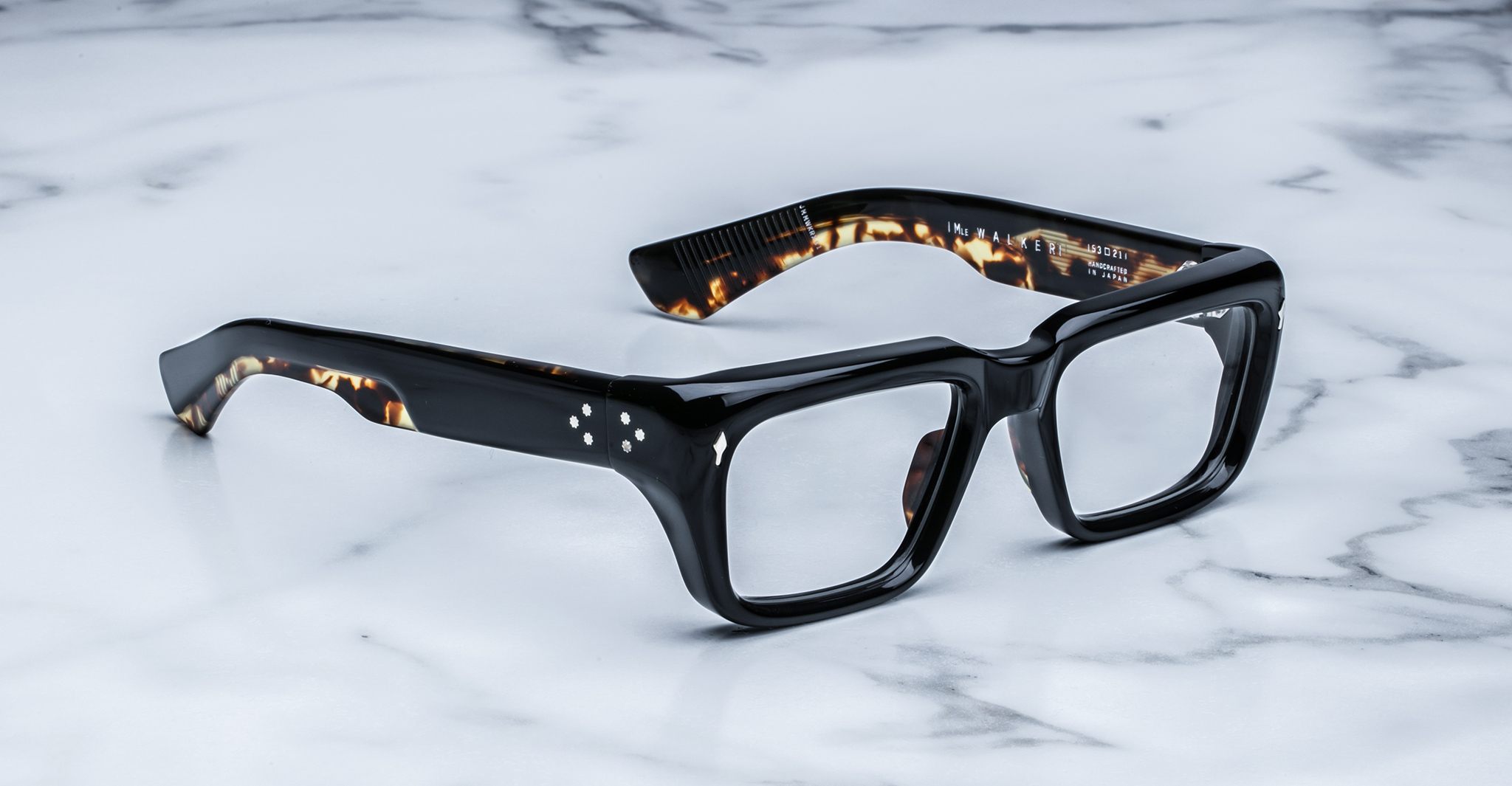 Thick, square-framed black eyeglasses with tortoiseshell inner details rest on a white marble surface. Four small white dots are visible on the temple of the frame.