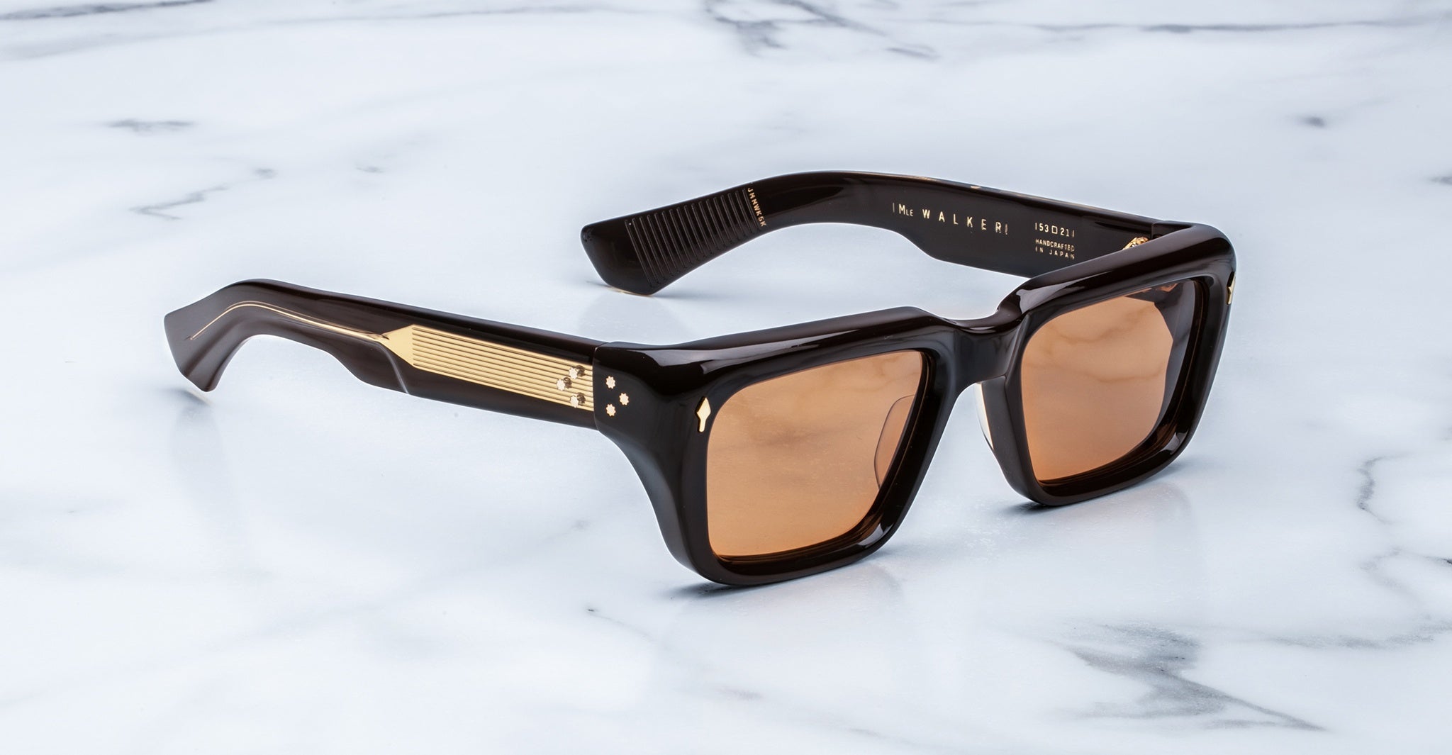 Black rectangular sunglasses with thick frames and brown lenses, featuring gold accents on the temples, displayed on a white marble surface.