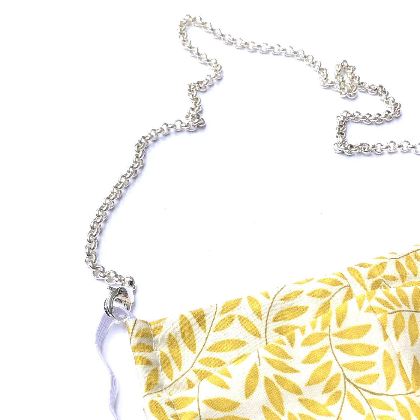 The Rolo Necklace Chain for Face Masks is attached to a corner of yellow and white leaf-patterned fabric, curving stylishly across a white background as both a functional mask holder and trendy jewelry accessory.