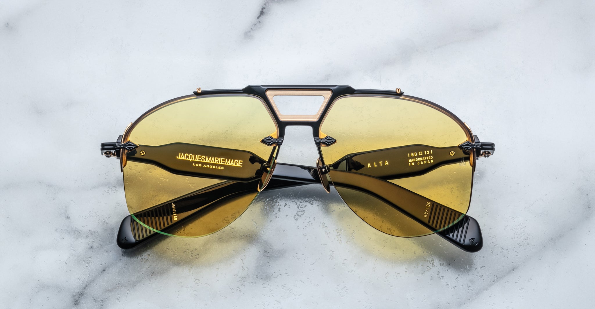 A pair of limited edition COMING SOON Jacques Marie Mage Alta aviator sunglasses with yellow-tinted lenses and black-and-gold frames rests on a white marble surface. The inside arms are labeled “Jacques Marie Mage Los Angeles” and “ALTA 18D01.”.