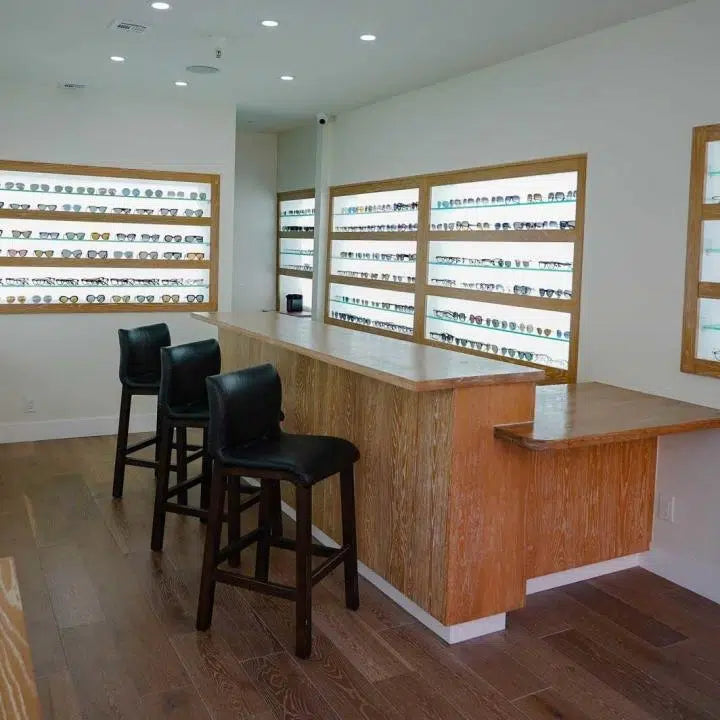 A modern eyewear store interior with wooden floors, a wooden counter with three black chairs, and walls lined with shelves displaying many pairs of eyeglasses and sunglasses.