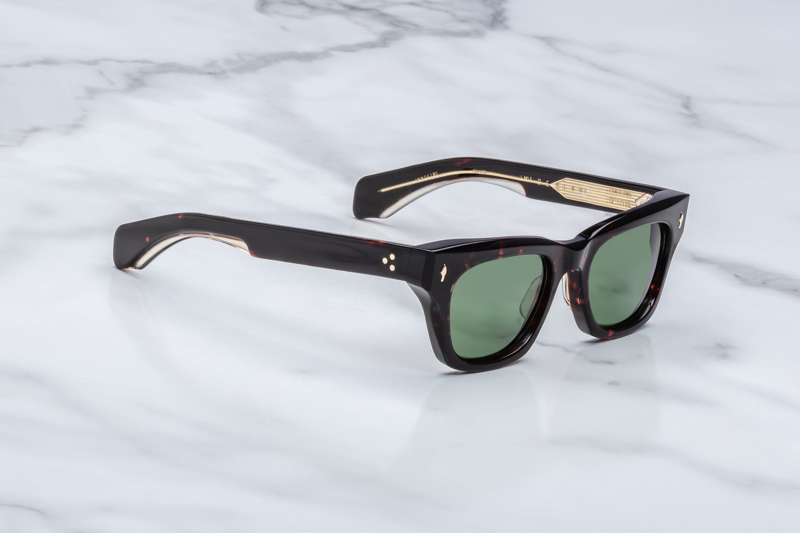 A pair of Jacques Marie Mage Dealan dark tortoiseshell square sunglasses with green lenses and gold accents rests on a white marble surface.