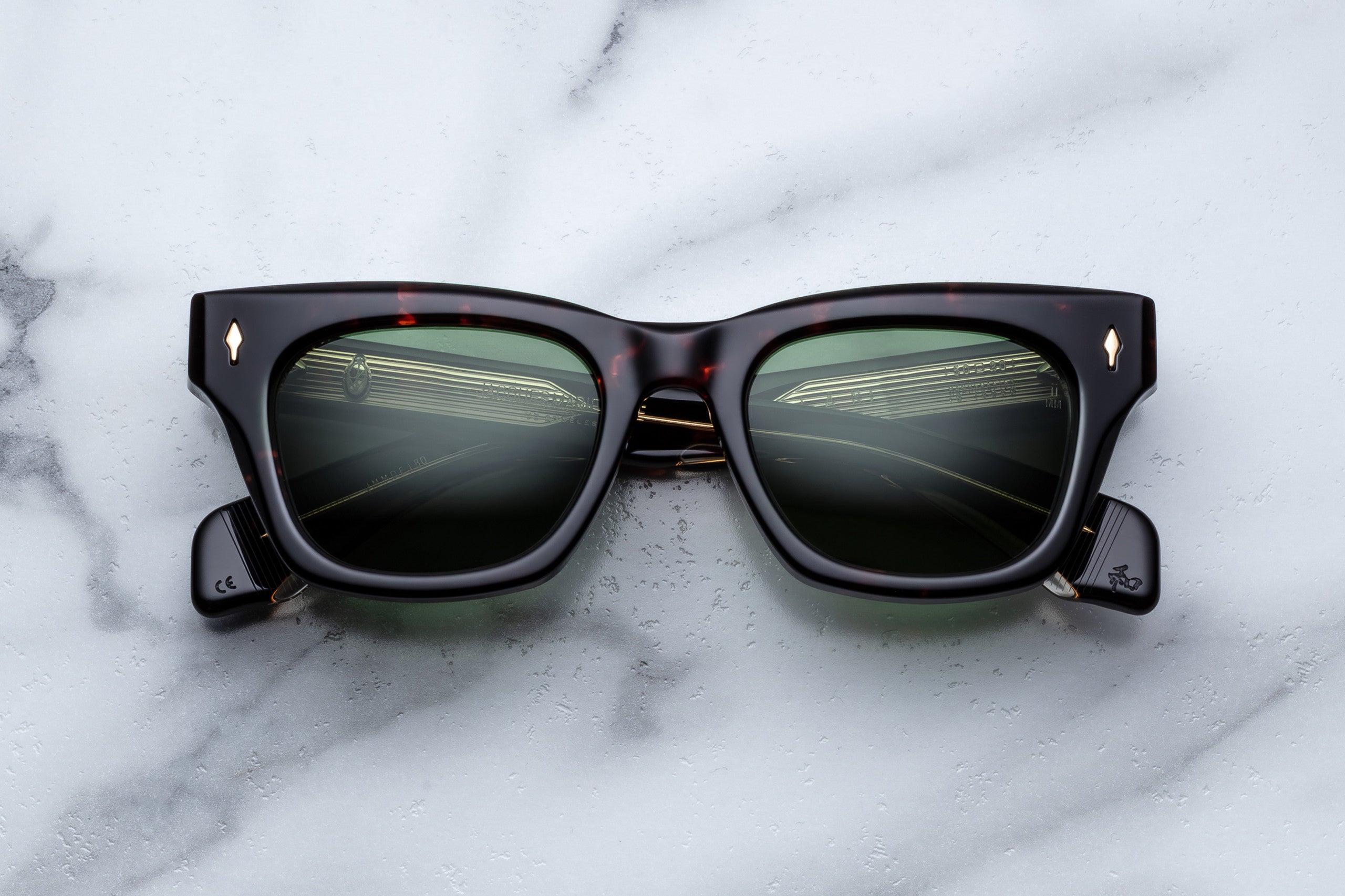 The Jacques Marie Mage Dealan sunglasses, featuring black frames and dark lenses, rest on a white marble surface. The lenses reflect a road with guardrails and a cloudy sky.