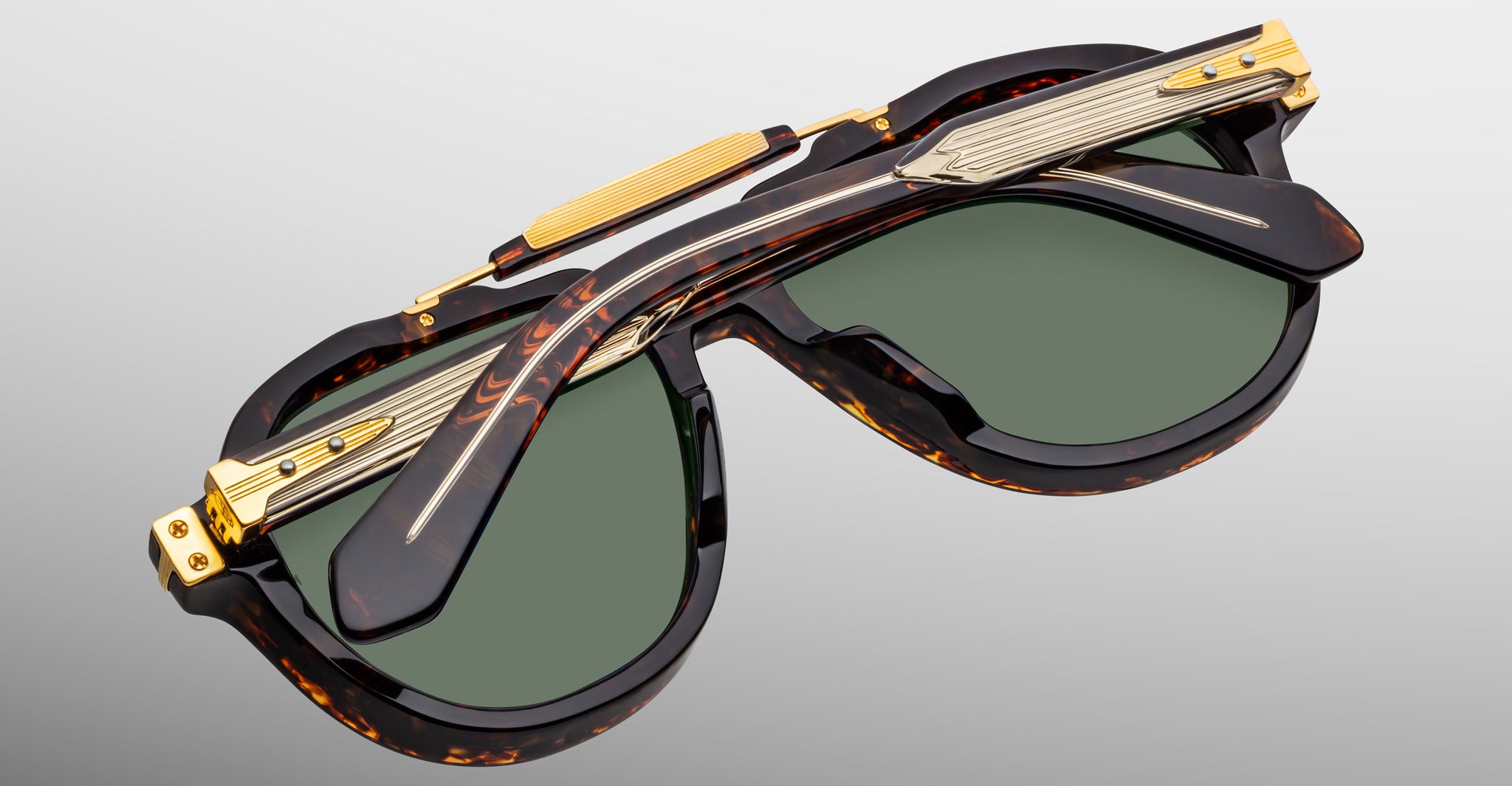 The Jacques Marie Mage 1991 luxury sunglasses feature green lenses, tortoiseshell-patterned frames, and gold-metal accents, shown folded on a gray surface.