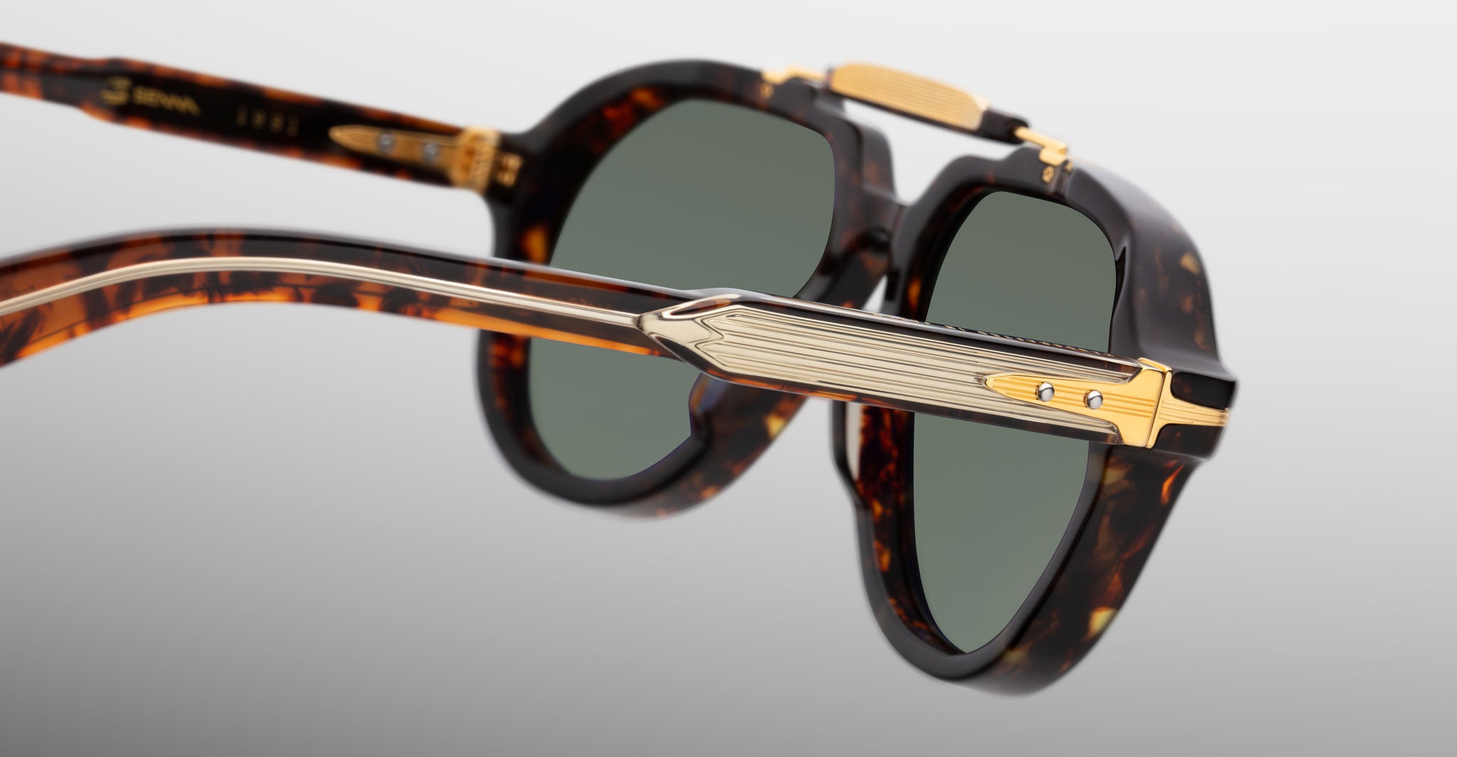 Close-up view of Jacques Marie Mage 1991 sunglasses in tortoiseshell with gold and textured metallic arm accents, featuring dark green tinted lenses, set against a light gray background.