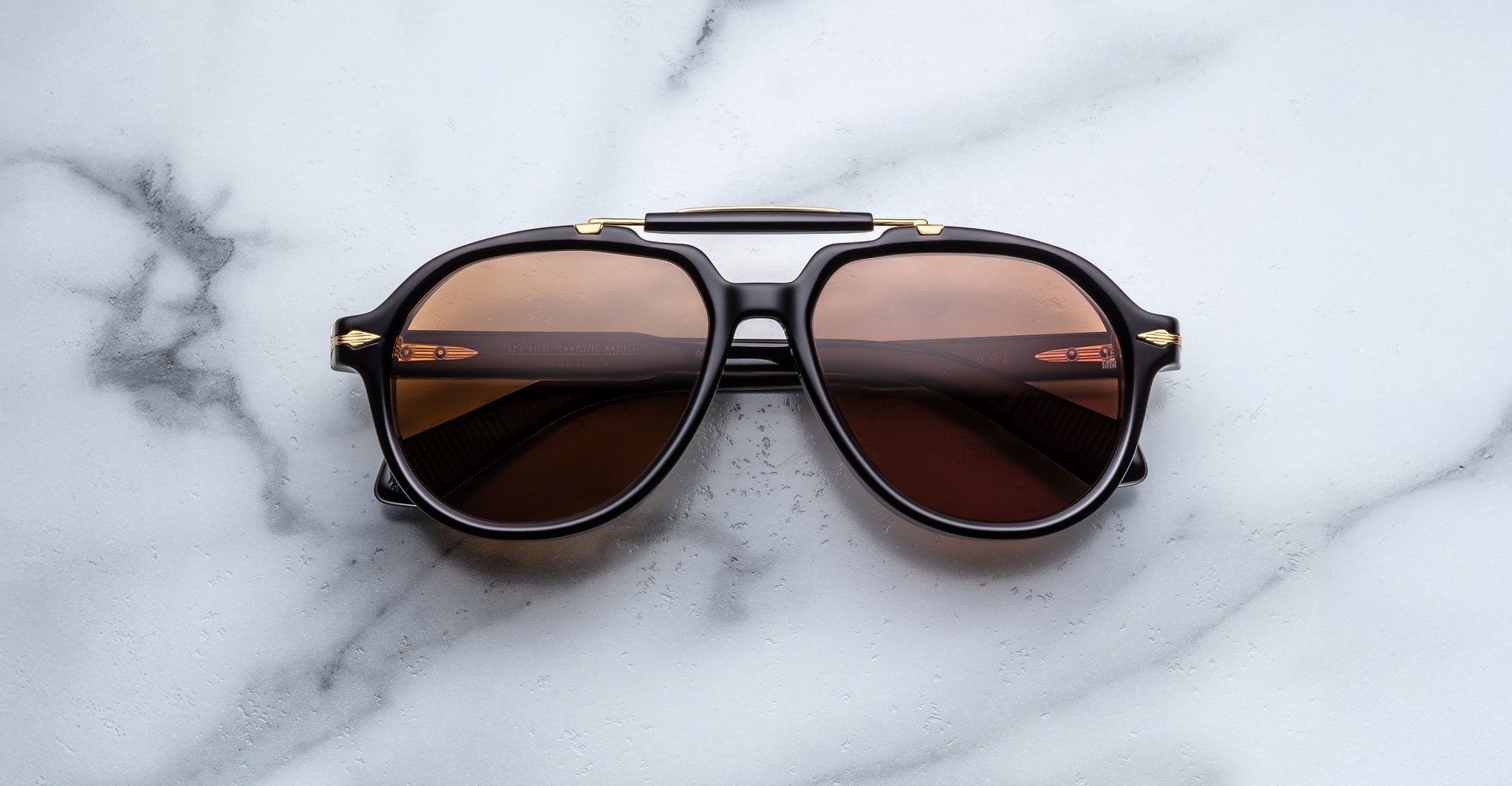 A pair of Jacques Marie Mage 1991 black sunglasses with brown-tinted lenses rests on a white marble surface, reflecting a modern railing and soft light in the lenses.