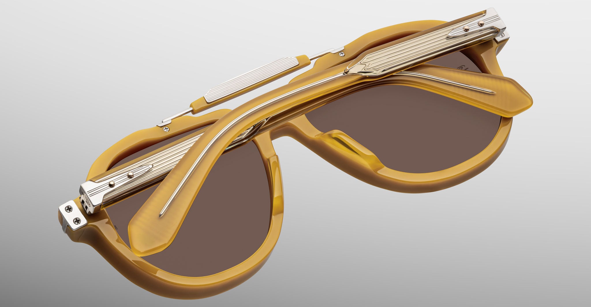 The Jacques Marie Mage 1991 sunglasses, featuring brown and gold frames with a unique folding bridge hinge and metallic accents, are displayed against a light gray background.