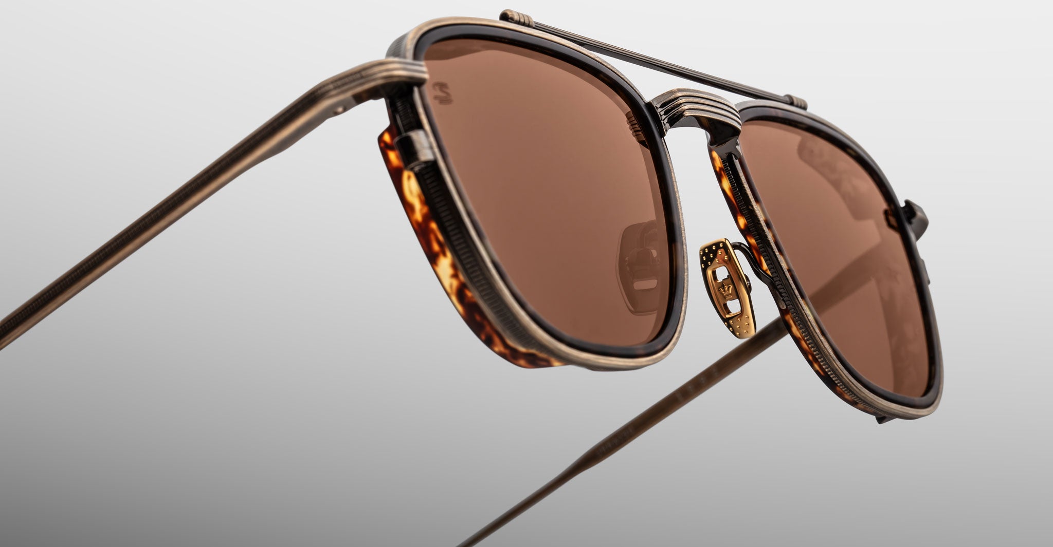 Close-up of Jacques Marie Mage 1992 sunglasses featuring brown-tinted lenses, a thin gold-toned metal frame, and tortoiseshell detailing around the lenses, set against a light gradient background.