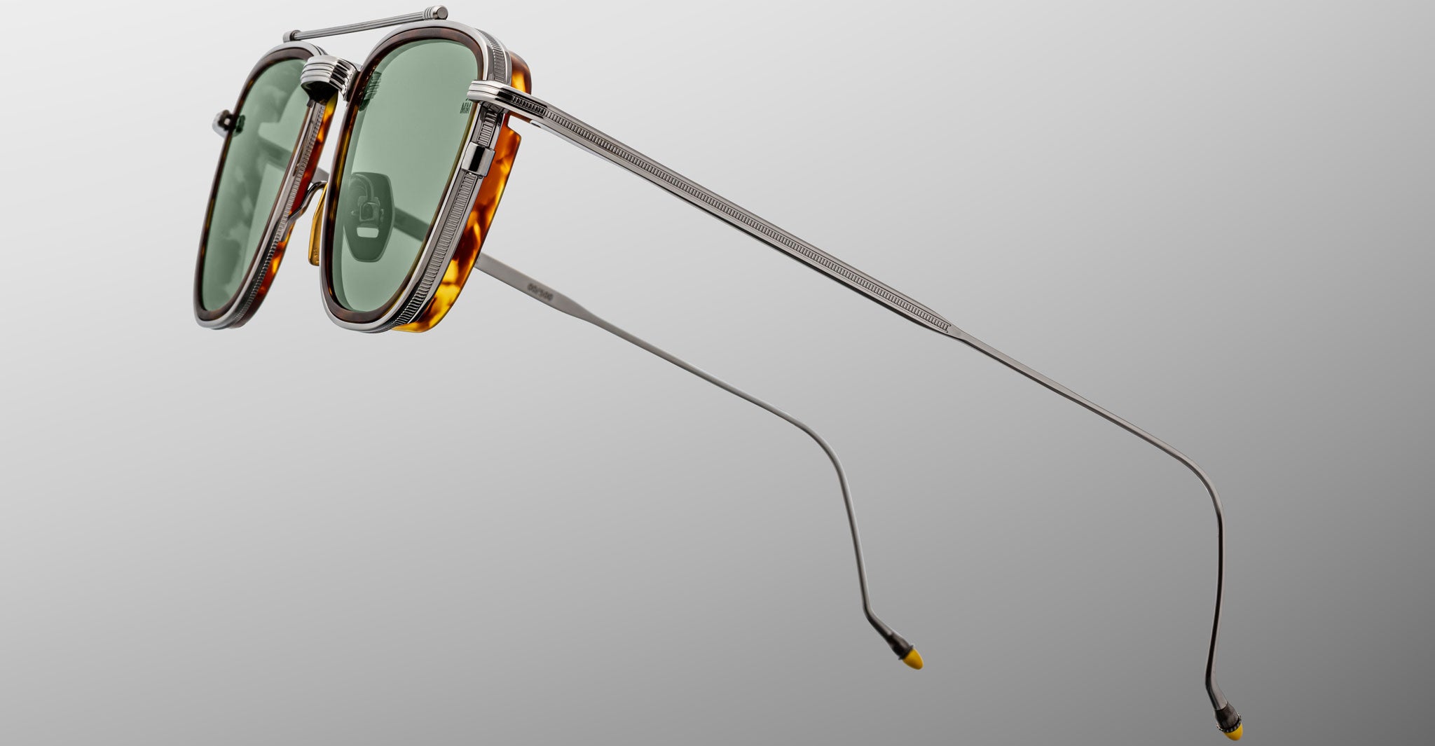 A close-up of Jacques Marie Mage 1992 sunglasses featuring green lenses, thin metallic arms, and tortoiseshell frames against a light gray background. The sunglasses are angled to showcase both the side profile and lens details.