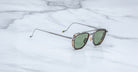 The Jacques Marie Mage 1992 sunglasses, featuring green lenses, thin silver metal arms, and a tortoiseshell frame, rest on a white marble surface with grey veining.
