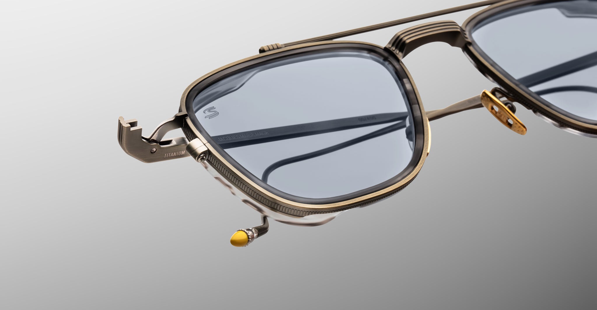 A close-up of Jacques Marie Mage 1992 sunglasses with stylish metallic frames, blue-tinted lenses, a tool-inspired temple detail, and a yellow tip accent, set against a gradient gray background.