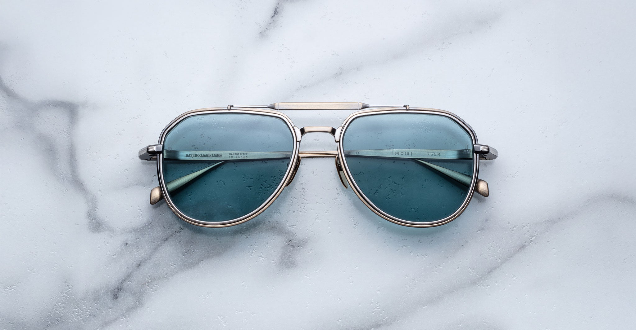The Jacques Marie Mage 755M aviator sunglasses, featuring blue-tinted lenses and a silver frame, rest on a white marble surface.