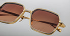 Close-up of Jacques Marie Mage Admiral sunglasses featuring stylish brown-tinted square lenses and gold metal frames, displayed against a light gray background.