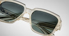 Close-up of Jacques Marie Mage Alfred sunglasses with clear frames, dark tinted lenses, and intricate metal detailing with branding on the inner arms, set against a neutral background.