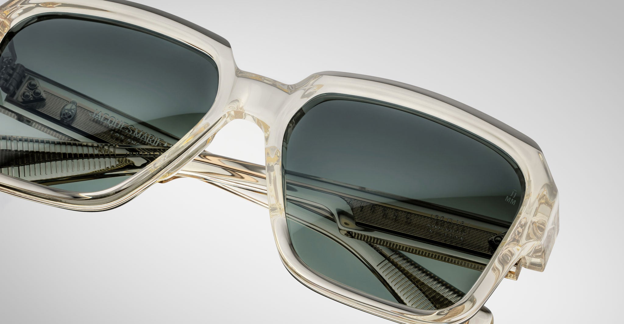 Close-up of Jacques Marie Mage Alfred sunglasses with clear frames, dark tinted lenses, and intricate metal detailing with branding on the inner arms, set against a neutral background.