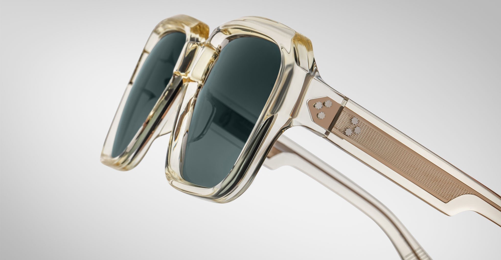 Close-up of Jacques Marie Mage Alfred sunglasses: stylish translucent square frames in thick beige, dark lenses, and metallic accents with small star details on the arms. Set against a plain light gray background.