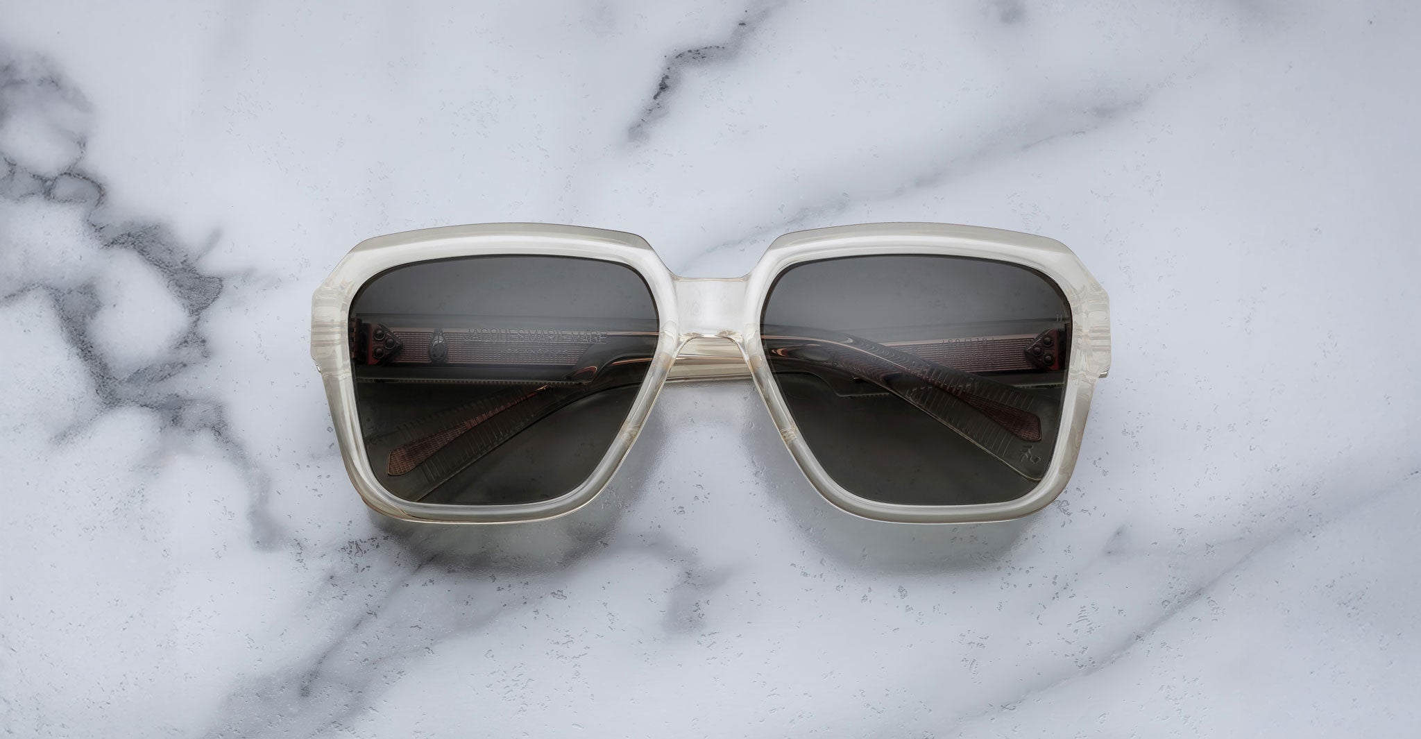 The Jacques Marie Mage Alfred sunglasses, featuring oversized square frames with translucent white rims and dark lenses, rest on a white marble surface veined with grey.
