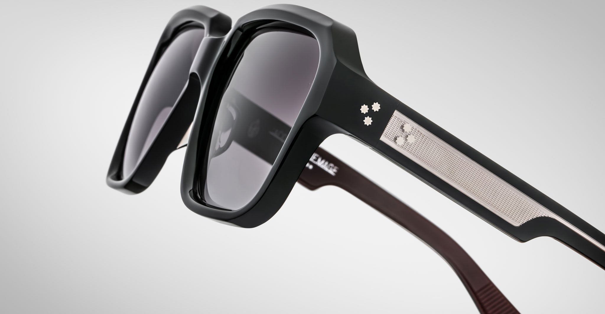 Close-up of Jacques Marie Mage Alfred sunglasses: black rectangular frame with dark lenses, metal accents, and two small decorative stars on the temple. Set against a plain light gray gradient background.