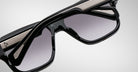 A close-up of Jacques Marie Mage Alfred sunglasses highlights their thick black frames, silver metal accents, and gradient gray lenses against a plain light background.