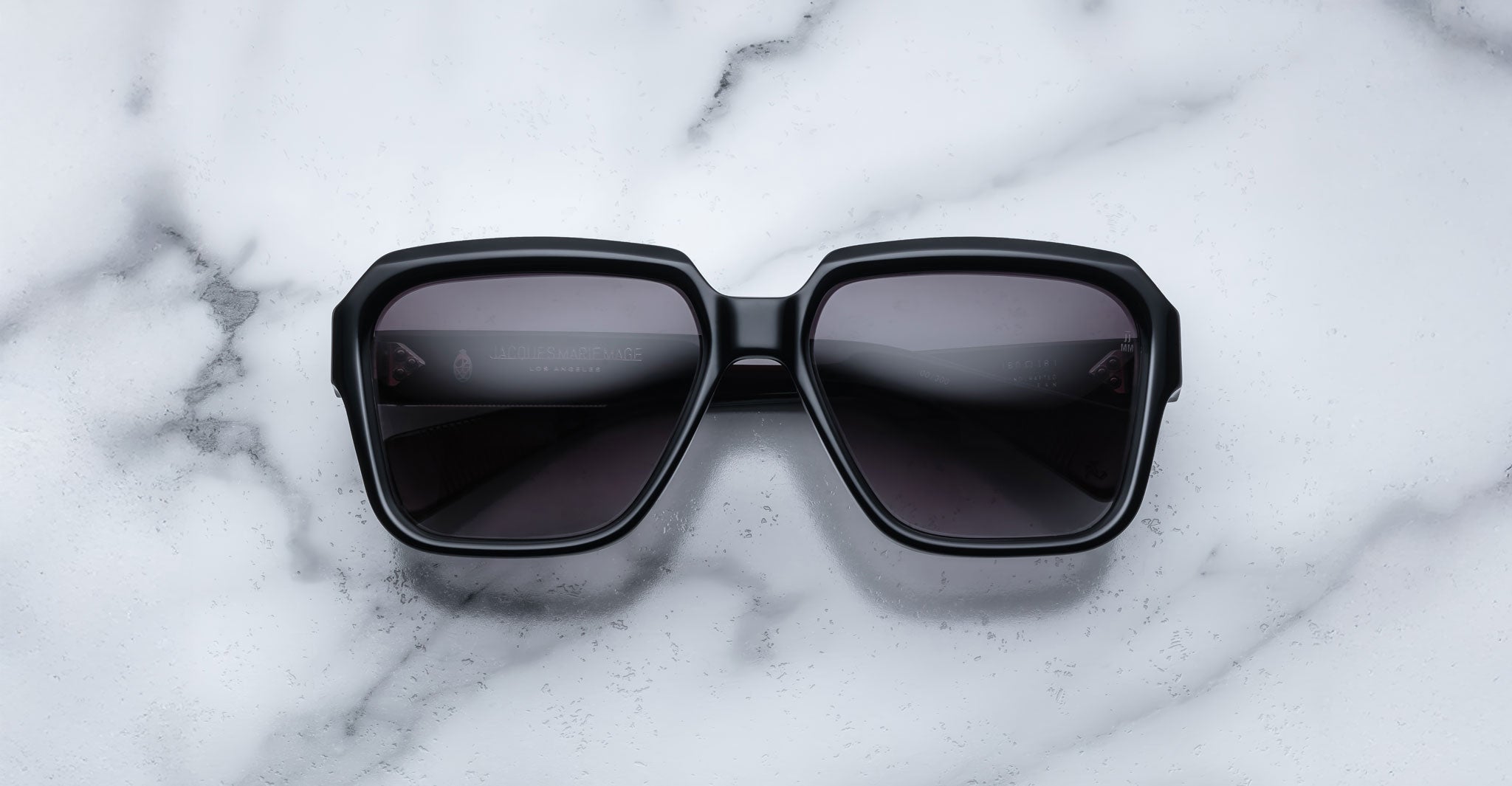 The Jacques Marie Mage Alfred, a pair of black square-framed sunglasses with dark lenses, is displayed on a white marble surface with gray veining.