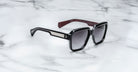 The Jacques Marie Mage Alfred sunglasses feature a black rectangular frame with silver detailing on the temples, maroon textured inner arms, and dark, slightly reflective lenses, displayed on a white marble surface.