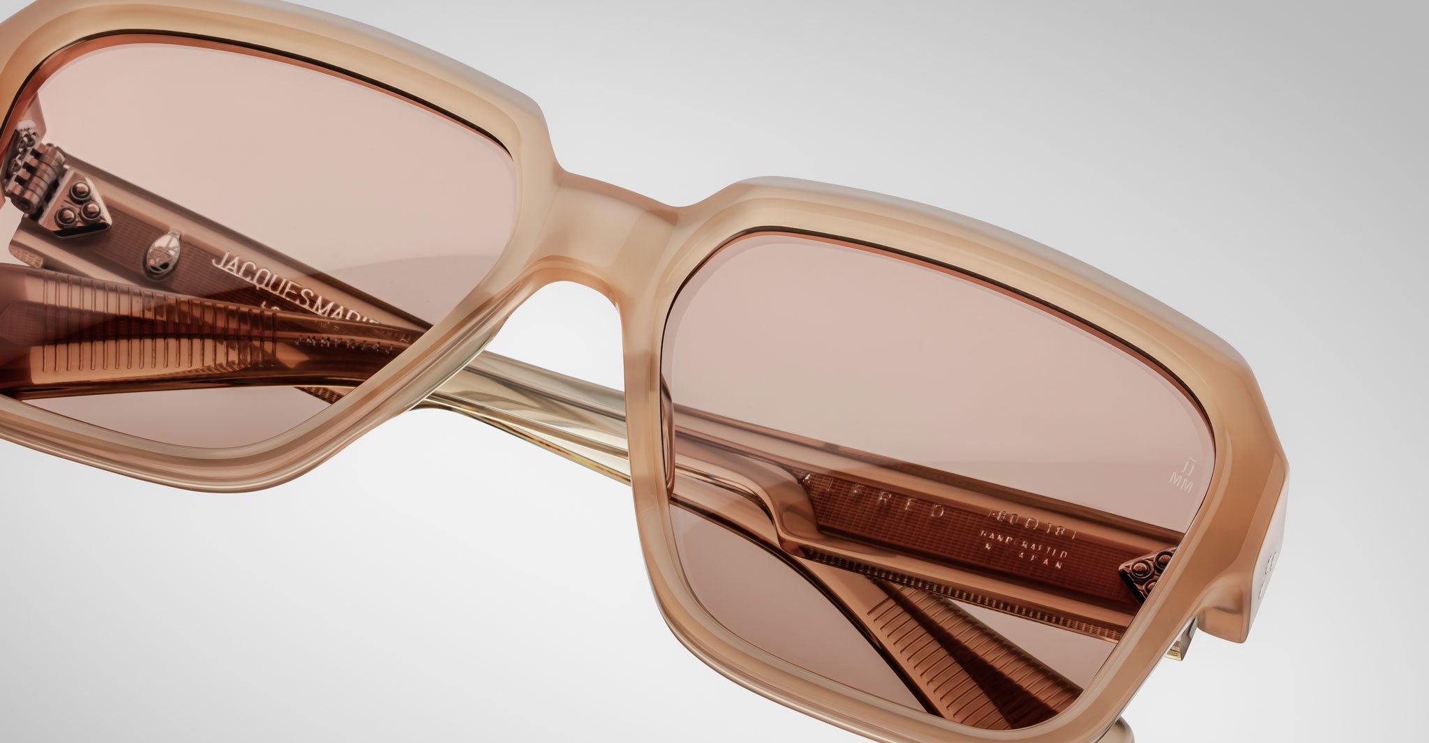 Close-up of Jacques Marie Mage Alfred sunglasses: beige, square-shaped frames with pink-tinted lenses and gold temple accents, set against a light gray background.
