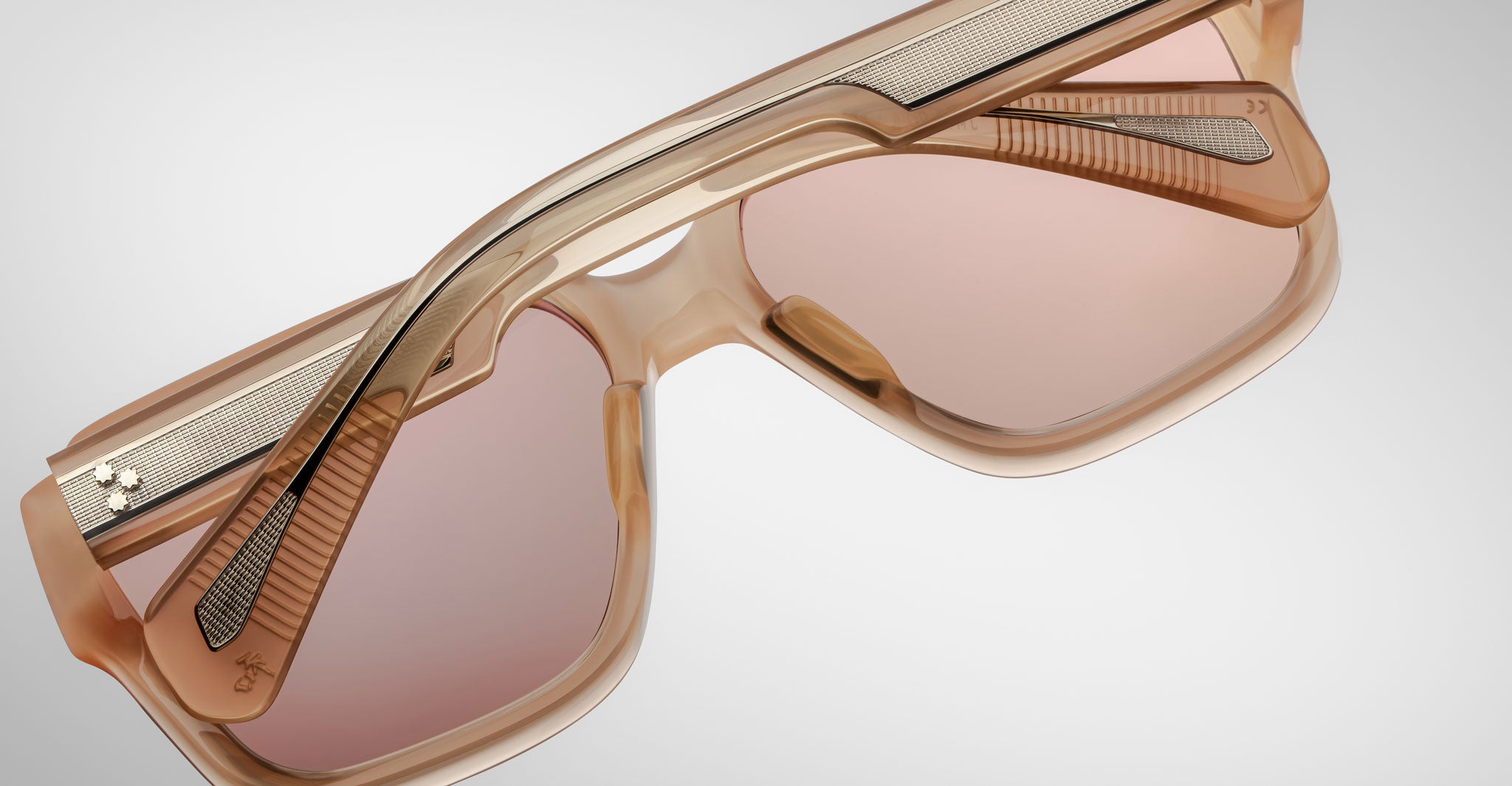 Close-up of Jacques Marie Mage Alfred sunglasses with pink-tinted lenses and a thick, light brown plastic frame featuring metallic accents and geometric temple and bridge details, set against a plain background.