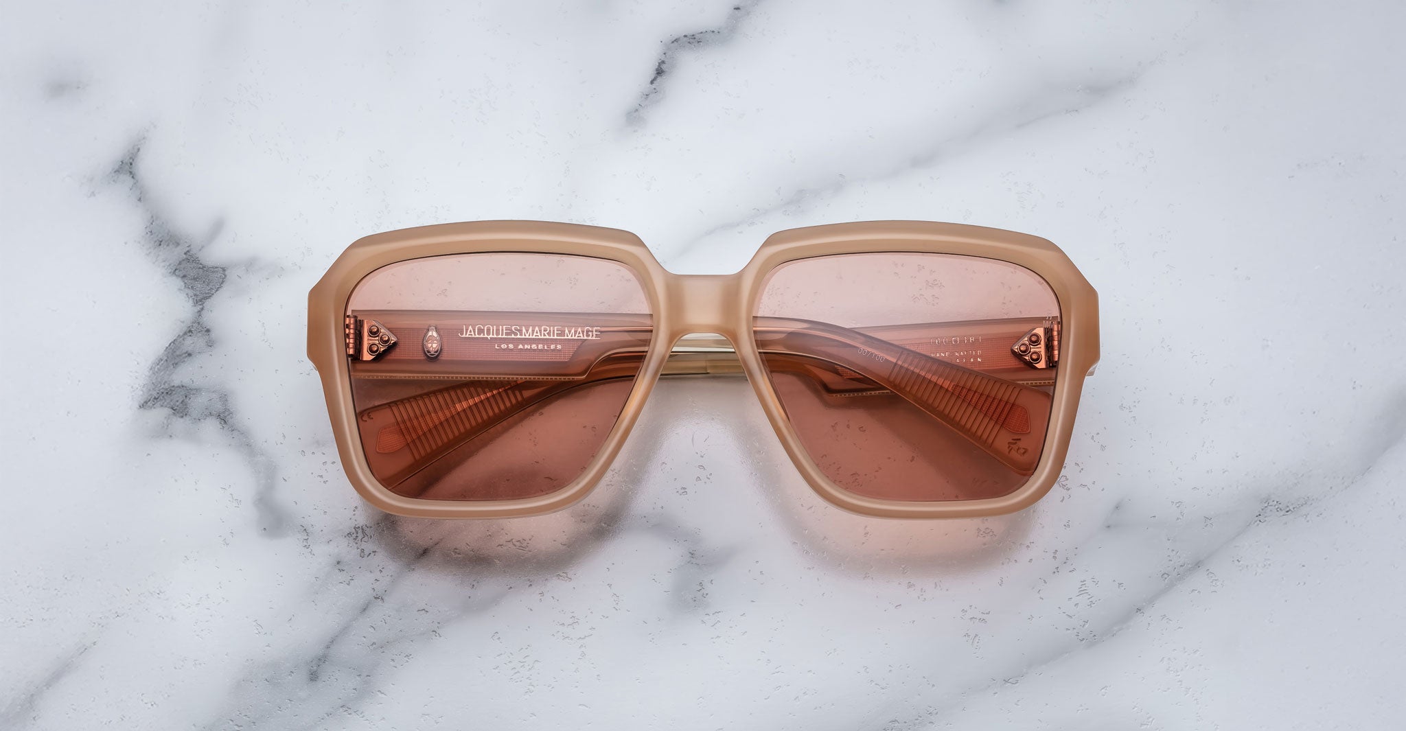 The Jacques Marie Mage Alfred sunglasses feature large, square frames in light brown with pink-tinted lenses, displayed on a white marble surface.