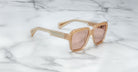 A pair of Jacques Marie Mage Alfred rectangular sunglasses with beige frames and light brown tinted lenses rests on a white marble surface.