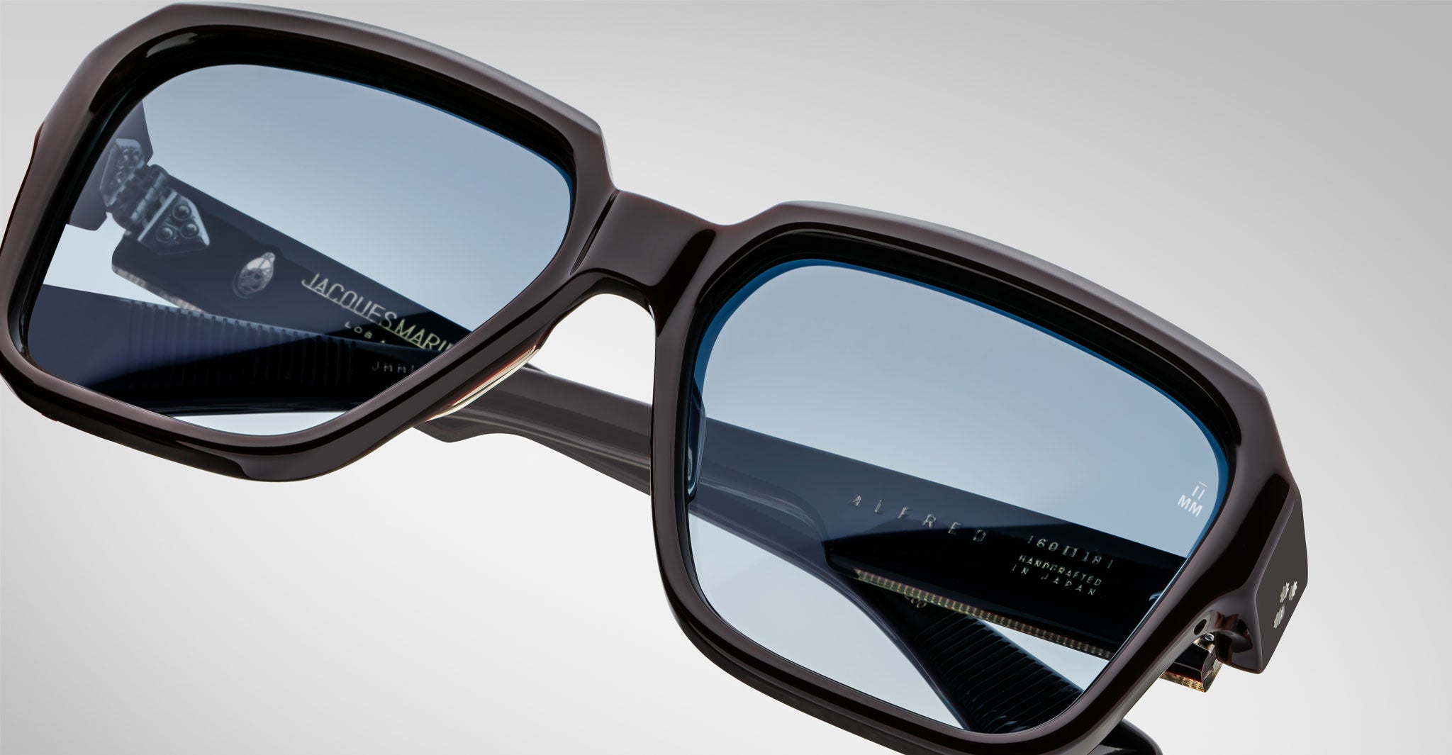 Close-up of Jacques Marie Mage Alfred rectangular eyeglasses featuring dark frames, light blue-tinted lenses, visible brand details, and intricate inner arm design against a plain neutral background.