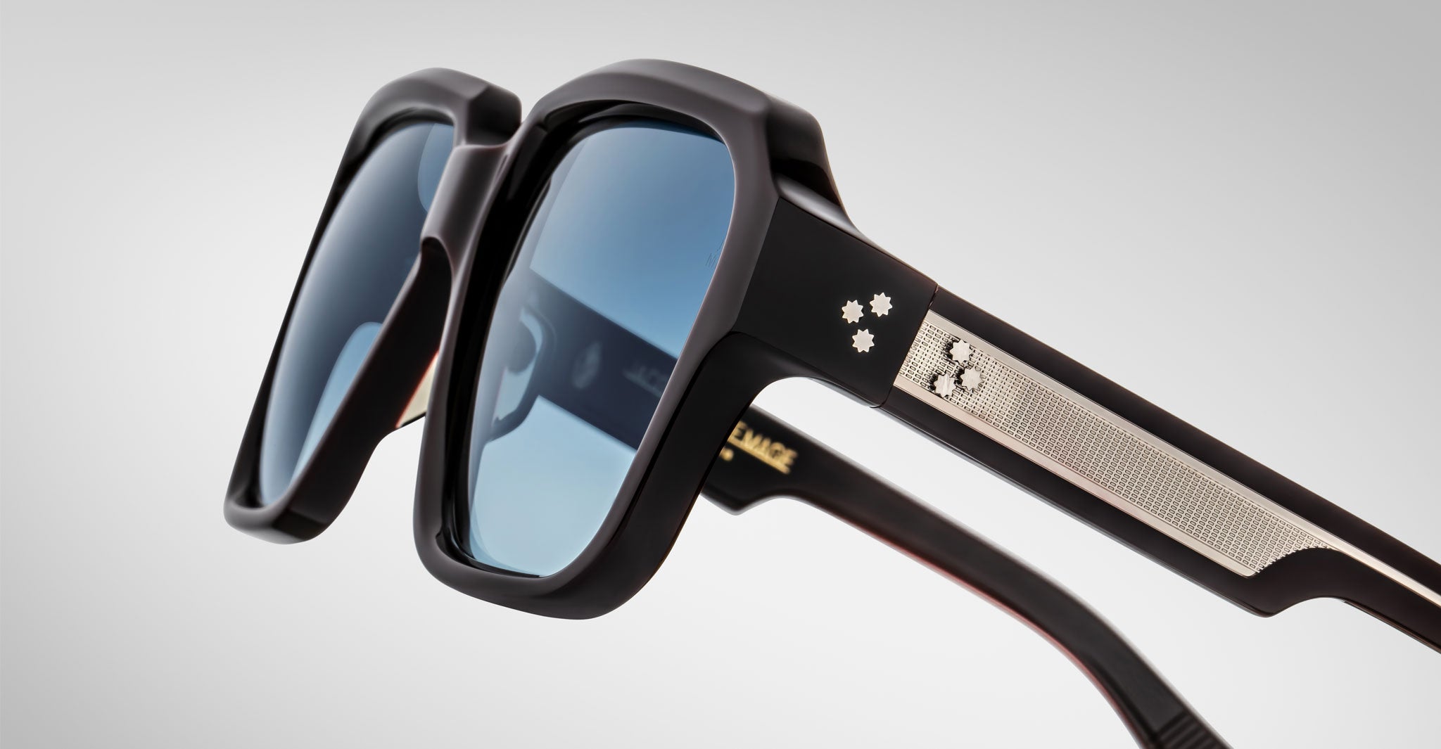 Close-up of Jacques Marie Mage Alfred sunglasses in matte black with blue-tinted lenses. The temple features a metallic detail adorned with three star-shaped accents and an engraved number 8.