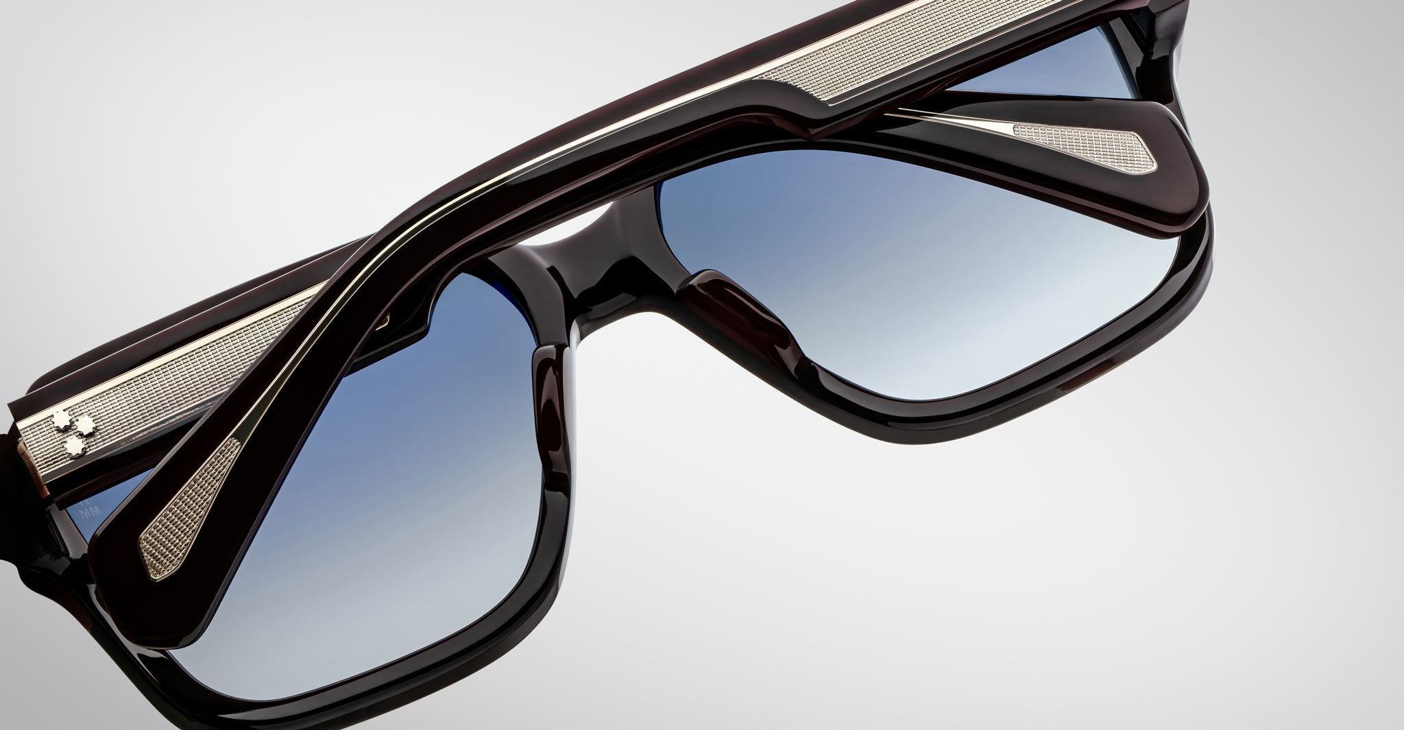 A close-up of Jacques Marie Mage Alfred sunglasses with black frames, blue gradient lenses, and metallic textured detailing on the arms and bridge, set against a light gray background.