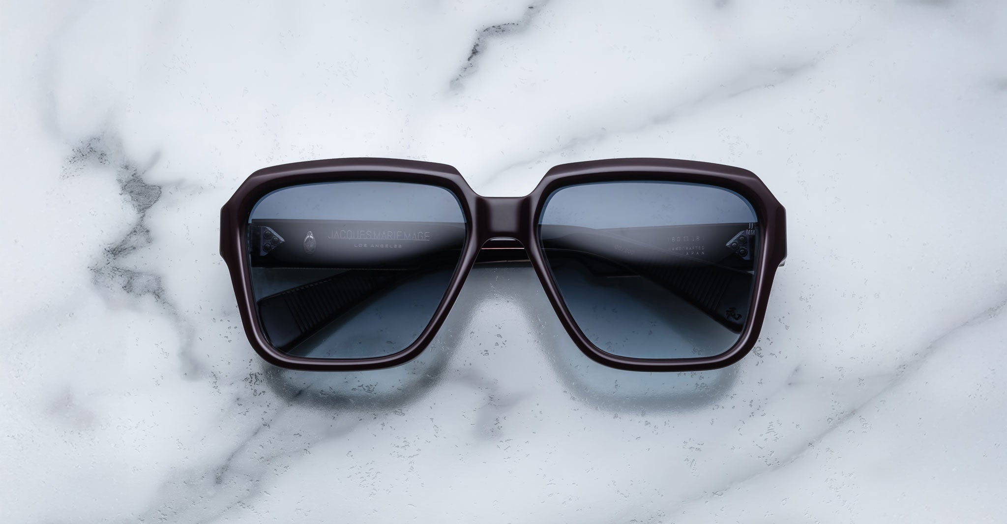 The Jacques Marie Mage Alfred sunglasses, featuring large square frames and dark lenses, are displayed on a white marble surface.