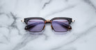 The Jacques Marie Mage Alin sunglasses feature dark purple lenses and a black and brown frame, displayed on a white marble surface.