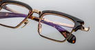 Close-up of Jacques Marie Mage Alin eyeglasses featuring a rectangular shape, dark tortoiseshell upper frame, gold accents, and clear lenses, resting on a light gray surface.