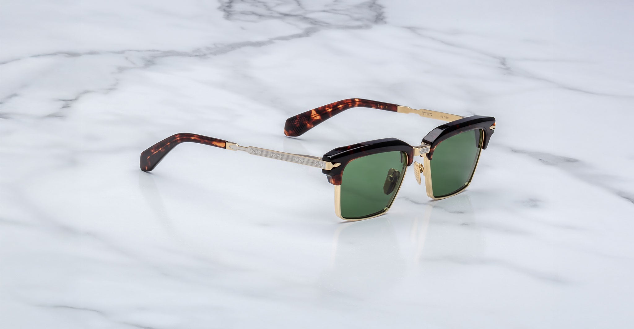 The Jacques Marie Mage Alin sunglasses feature green lenses, gold metal frames, tortoiseshell arms, and black accents, displayed on a white marble surface.