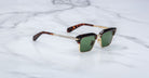 The Jacques Marie Mage Alin sunglasses feature green lenses, gold metal frames, tortoiseshell arms, and black accents, displayed on a white marble surface.