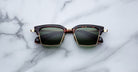 The Jacques Marie Mage Alin sunglasses, featuring tortoiseshell and gold frames with dark green lenses, rest on a white marble surface.