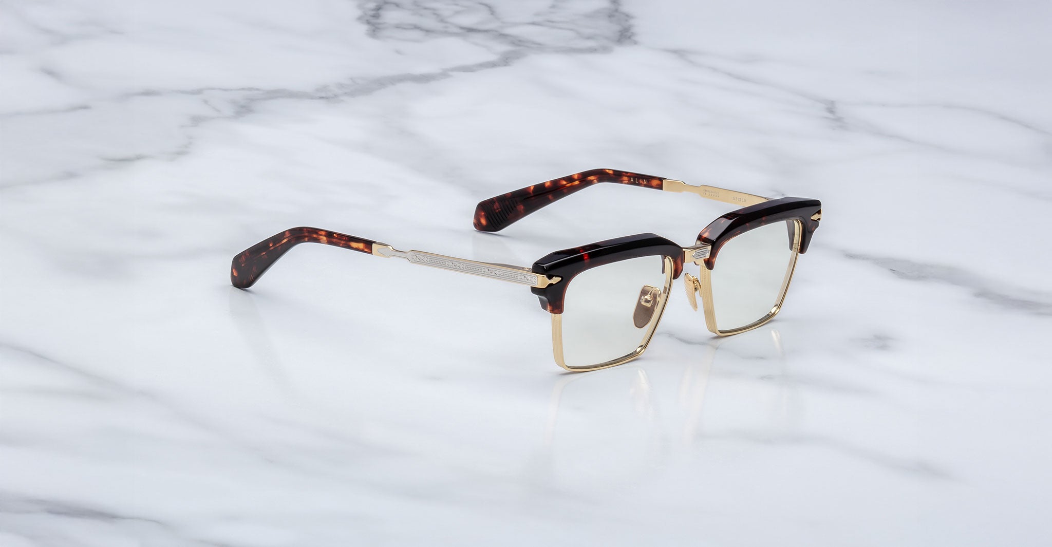 The Jacques Marie Mage Alin eyeglasses feature gold metal frames, tortoiseshell-patterned arms, clear rectangular lenses, and a browline detail, shown resting on a white marble surface.