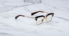 The Jacques Marie Mage Alin eyeglasses feature gold metal frames, tortoiseshell-patterned arms, clear rectangular lenses, and a browline detail, shown resting on a white marble surface.