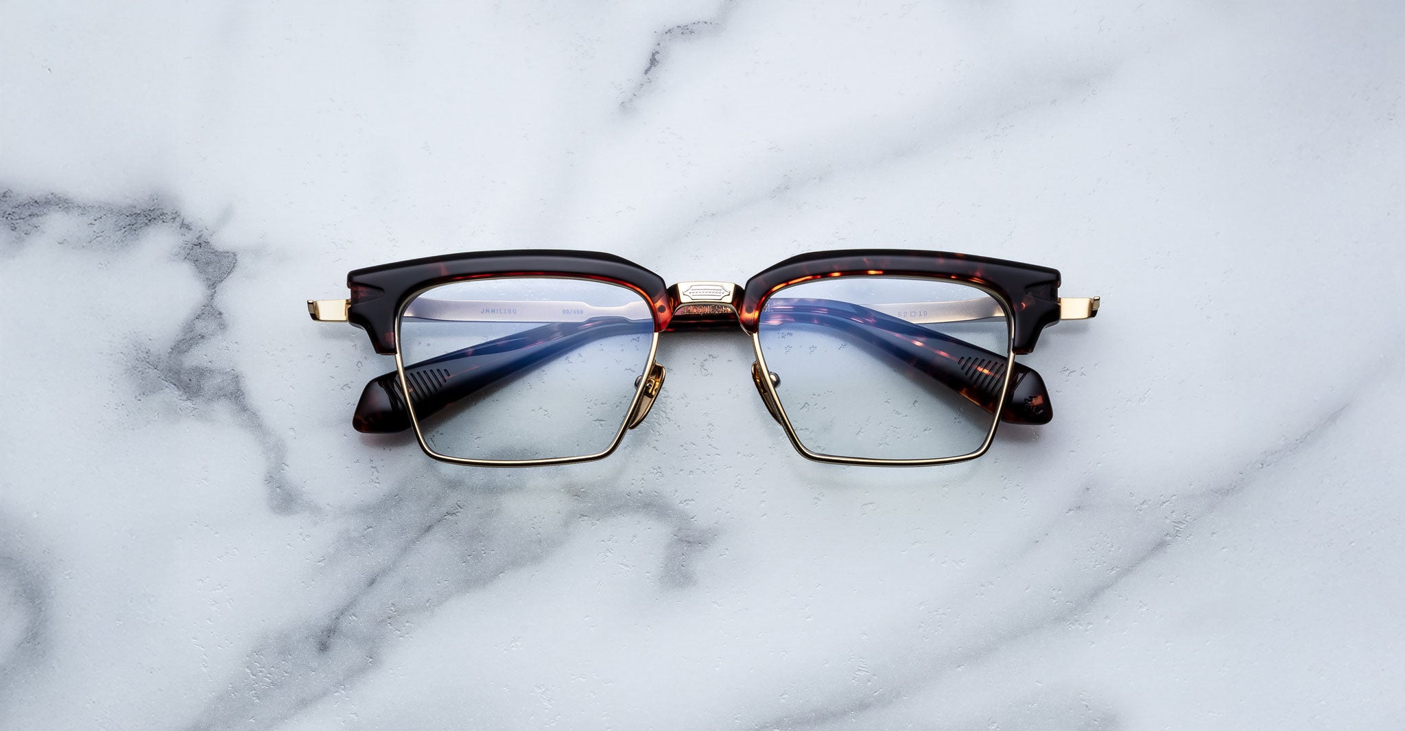 The Jacques Marie Mage Alin browline eyeglasses, featuring tortoiseshell frames and gold accents, are displayed on a white marble surface.