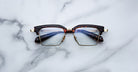 The Jacques Marie Mage Alin browline eyeglasses, featuring tortoiseshell frames and gold accents, are displayed on a white marble surface.