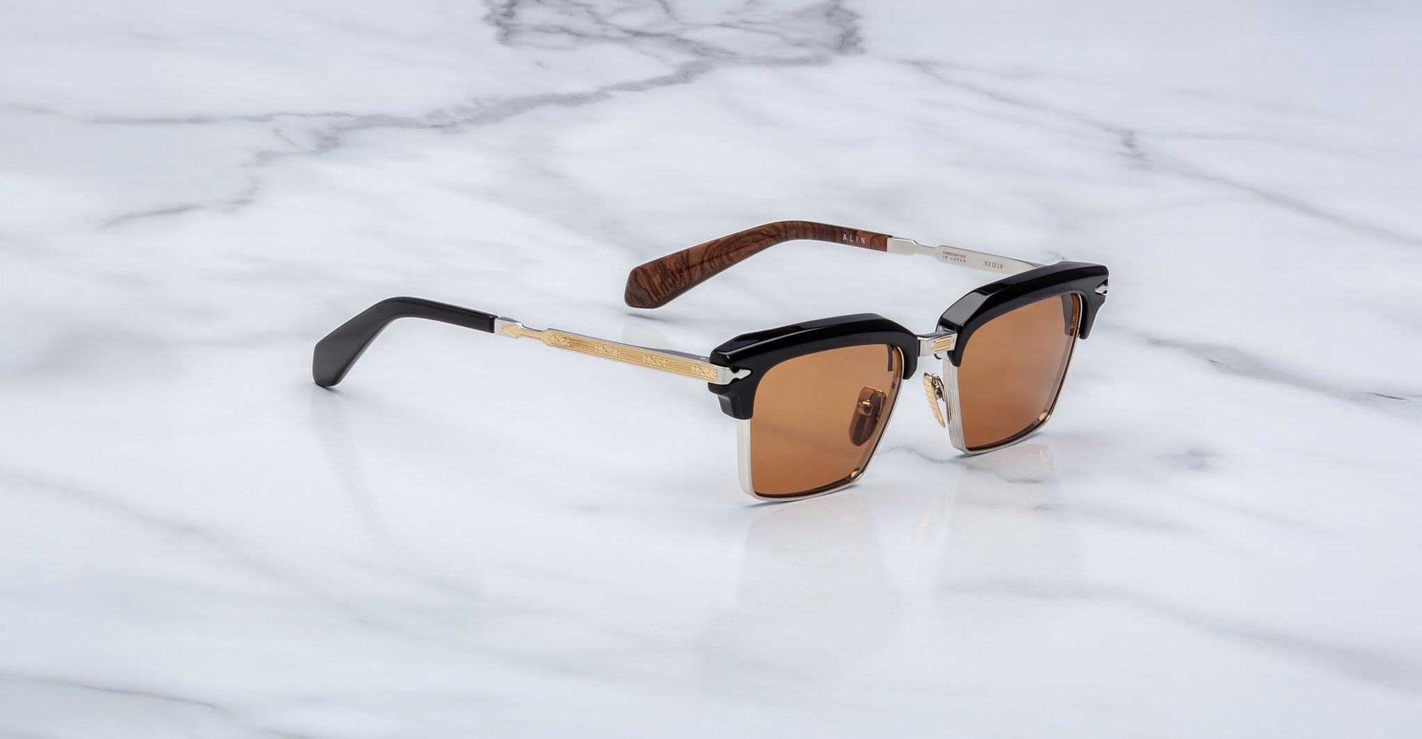 The Jacques Marie Mage Alin sunglasses feature brown lenses, black and gold frames, and wood-patterned temples, displayed on a white marble surface.