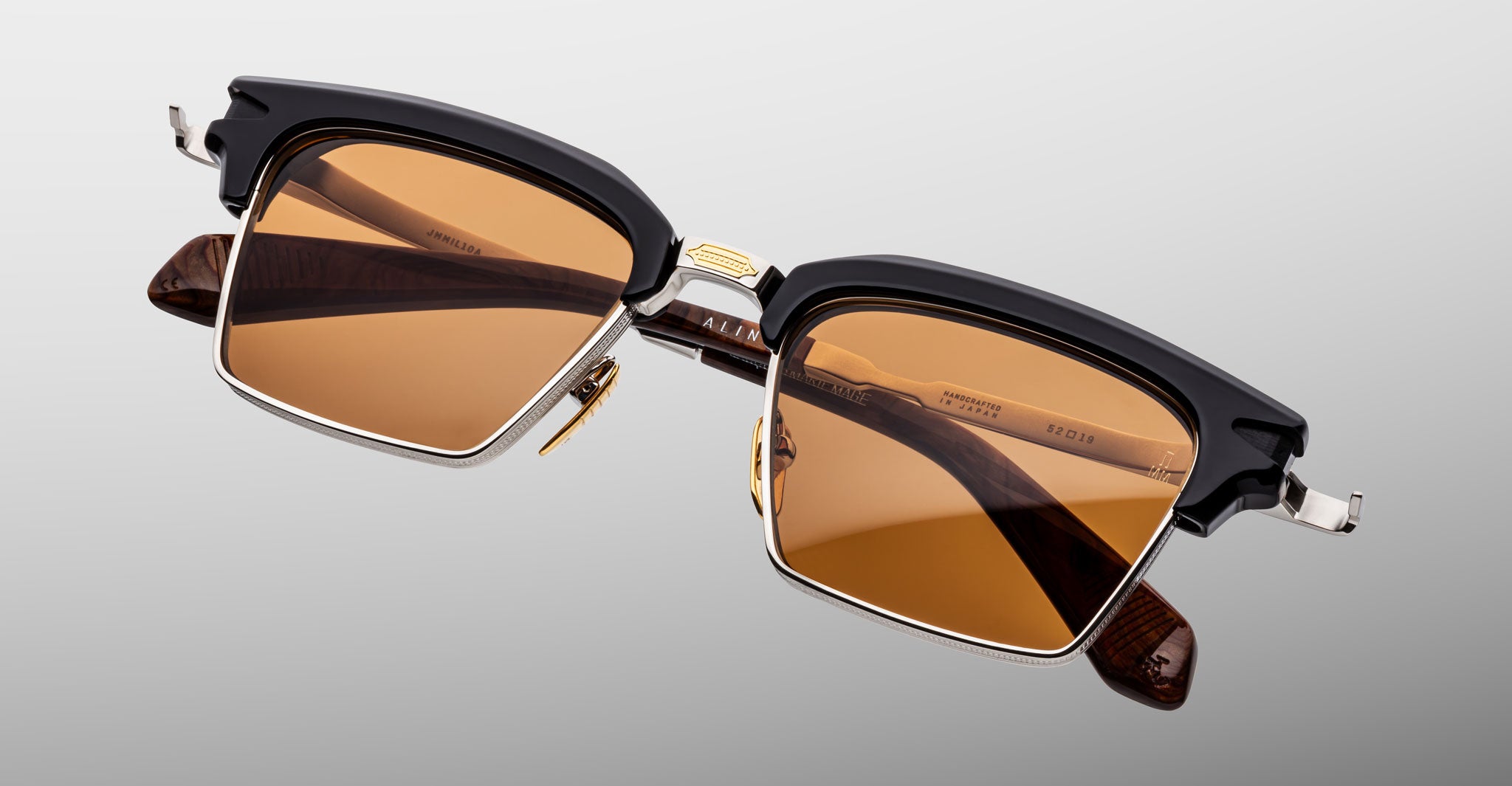 The Jacques Marie Mage Alin features rectangular sunglasses with brown-tinted lenses, black and gold frames, and tortoiseshell-patterned arms, shown on a light gray background.