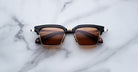 The Jacques Marie Mage Alin sunglasses, featuring black and gold rectangular frames with brown tinted lenses, are displayed on a white marble surface.