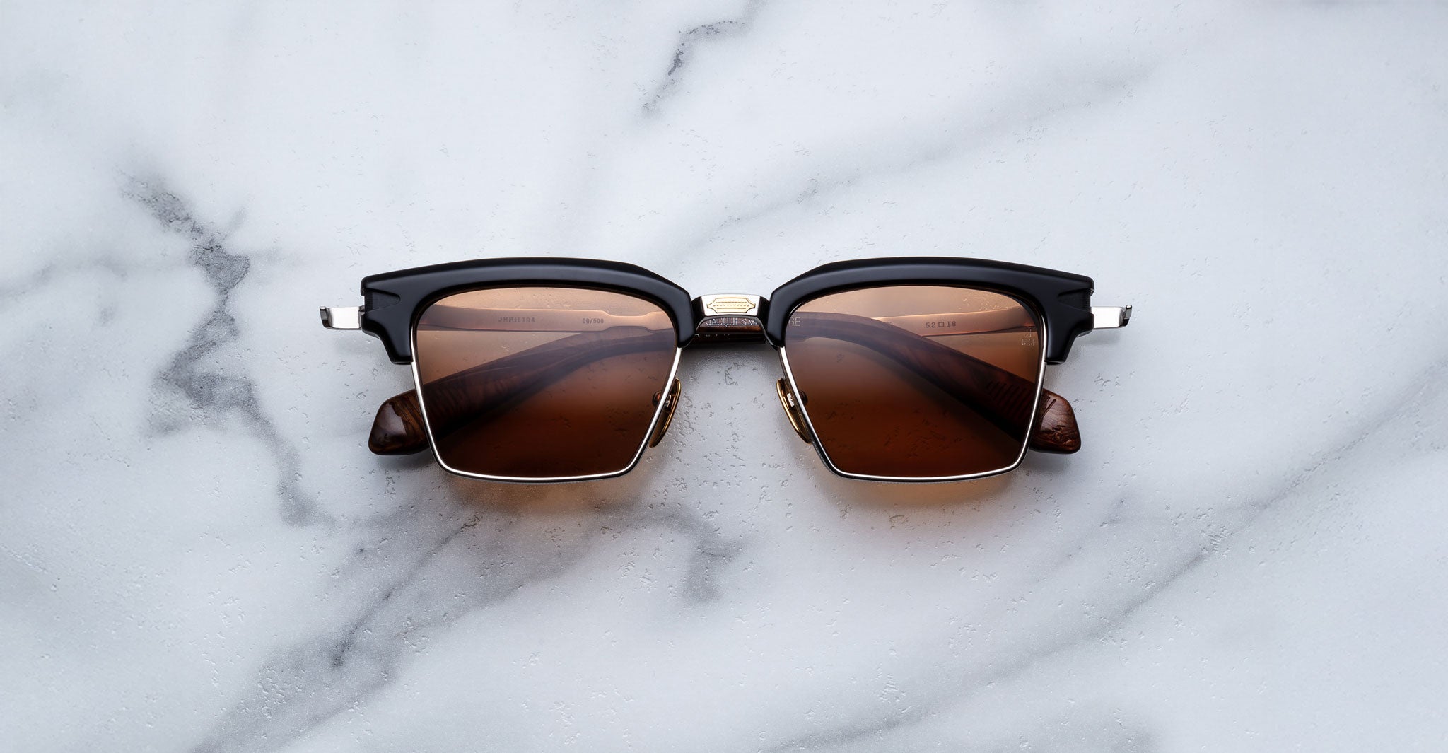 The Jacques Marie Mage Alin sunglasses, featuring black and gold rectangular frames with brown tinted lenses, are displayed on a white marble surface.